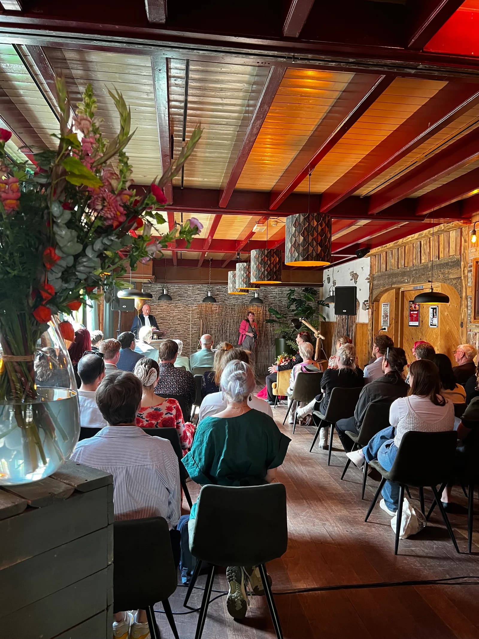 Volle ruimte tijdens een ceremonie, met een hoog bloemstuk op de voorgrond