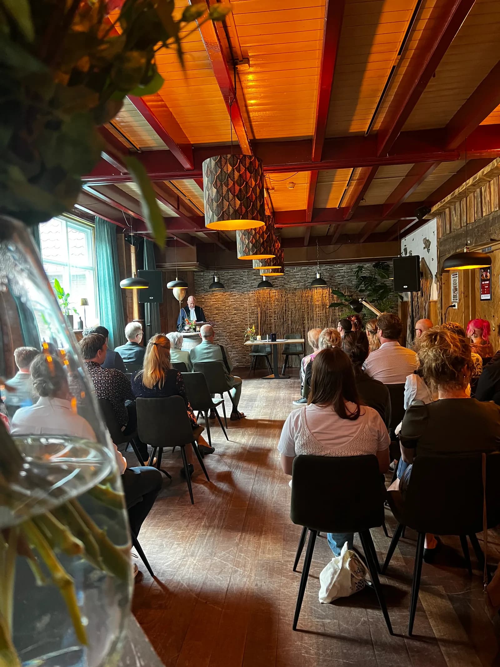 Gasten luisteren naar de spreker tijdens een ceremonie, gezien over een bloemstuk heen