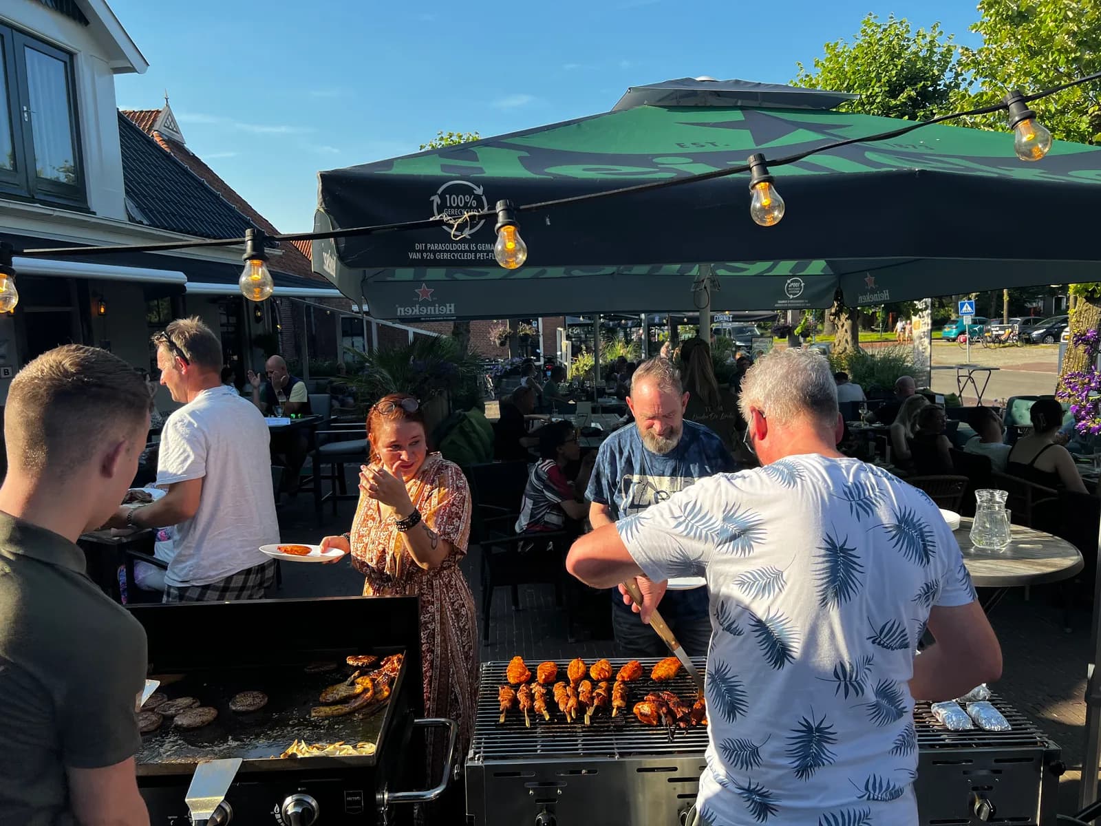Twee koks bakken hamburgers en spiesjes op de barbecue naast het terras, met gasten die hun bord laten vullen