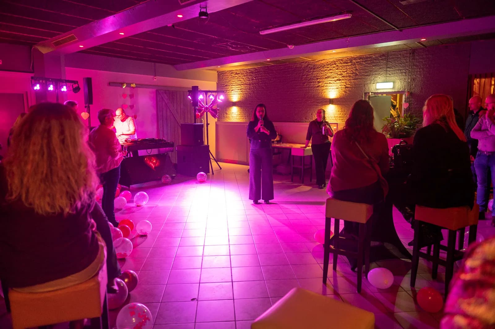 Zaal in paars licht met dj, gasten aan statafels en ballonnen op de vloer