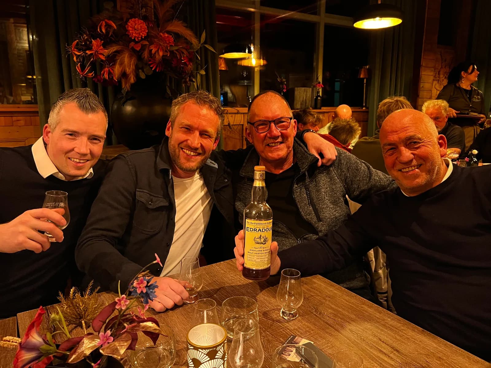 Vier lachende mannen aan een tafel in de gastrobar met een fles Edradour single malt whisky en proefglazen, in avondlicht