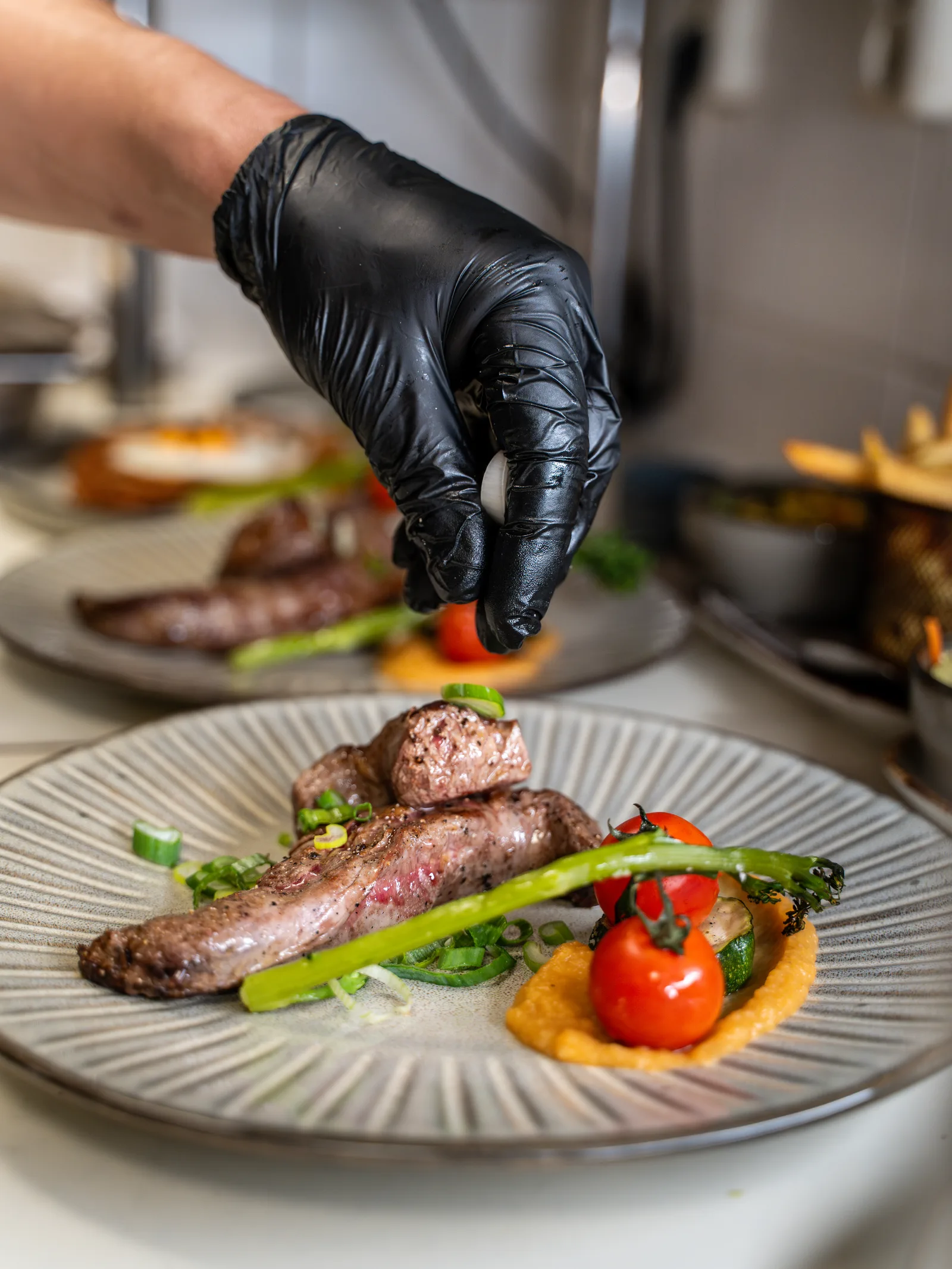Kok legt met een zwarte handschoen het laatste tomaatje op een bord met vlees, asperge en wortelcrème