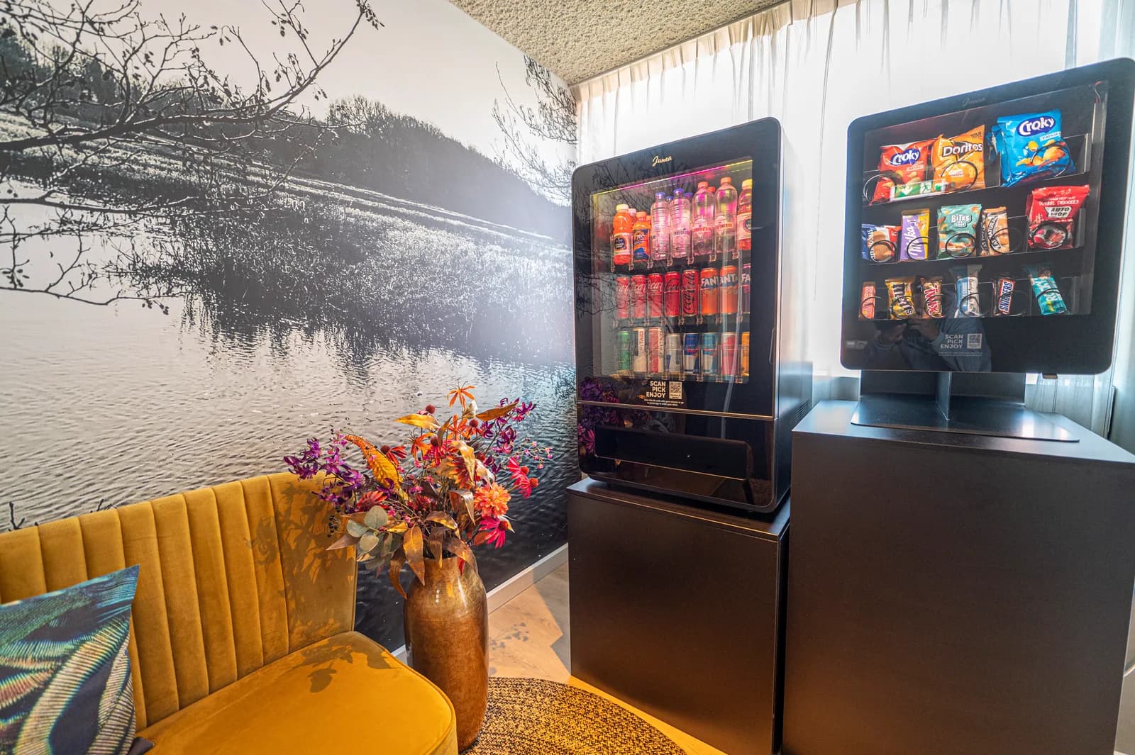 Zithoek op de hotelgang met okergele bank, fotobehang en twee automaten met drank en snacks
