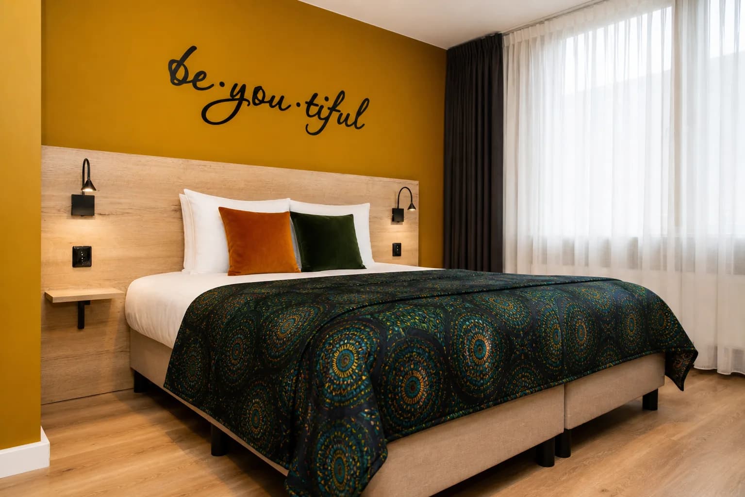 Hotelkamer met okergele wand, de muurtekst be.you.tiful en een tweepersoonsbed met een donkergroene sprei