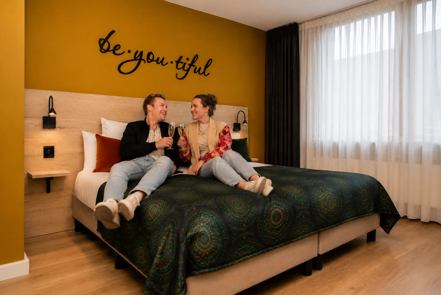 Stel proost met cava op het bed van een hotelkamer, tegen de okergele wand met de muurtekst be.you.tiful