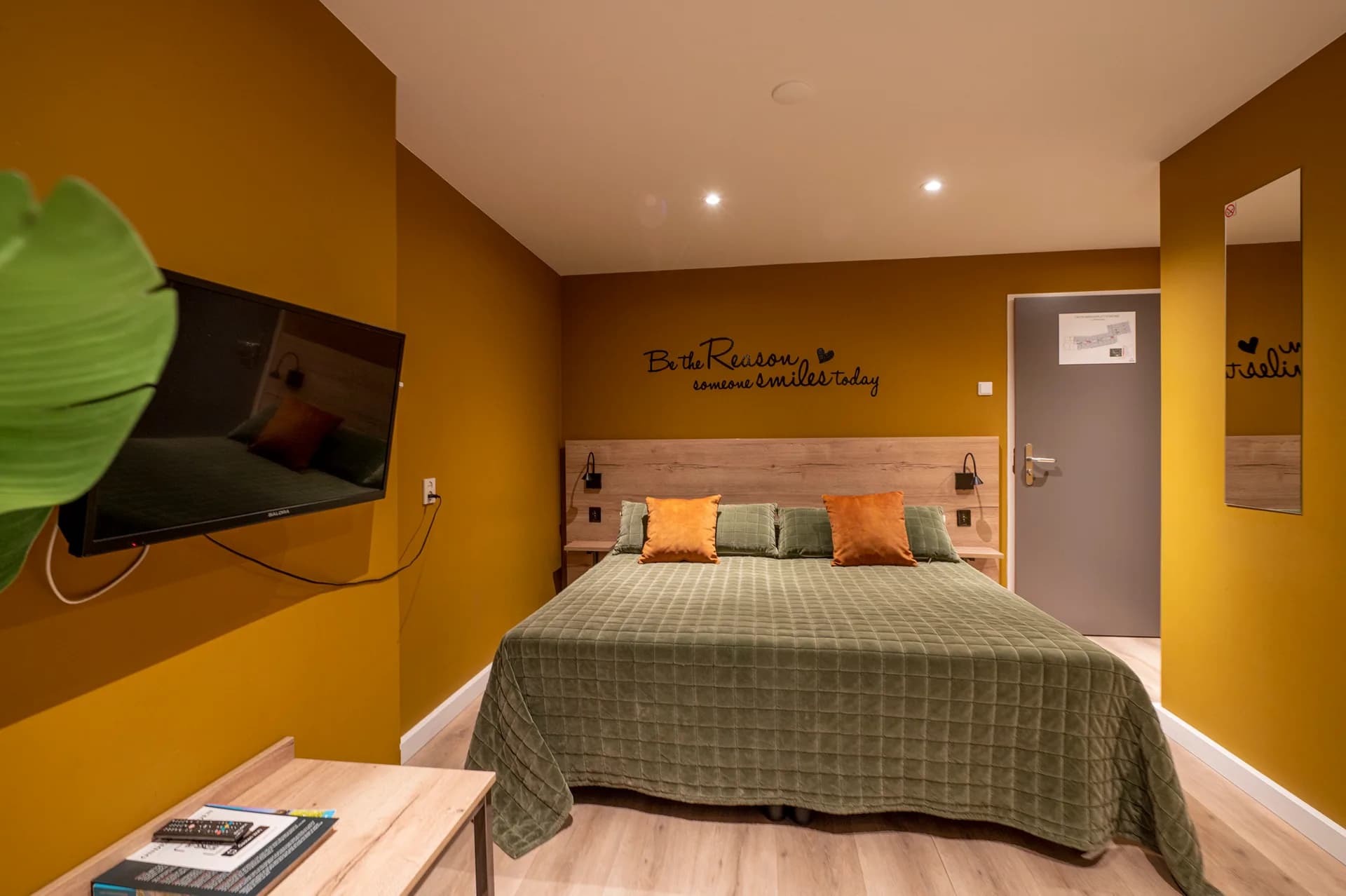 Hotelkamer met okergele wanden, tv, spiegel en de muurtekst be the reason someone smiles today