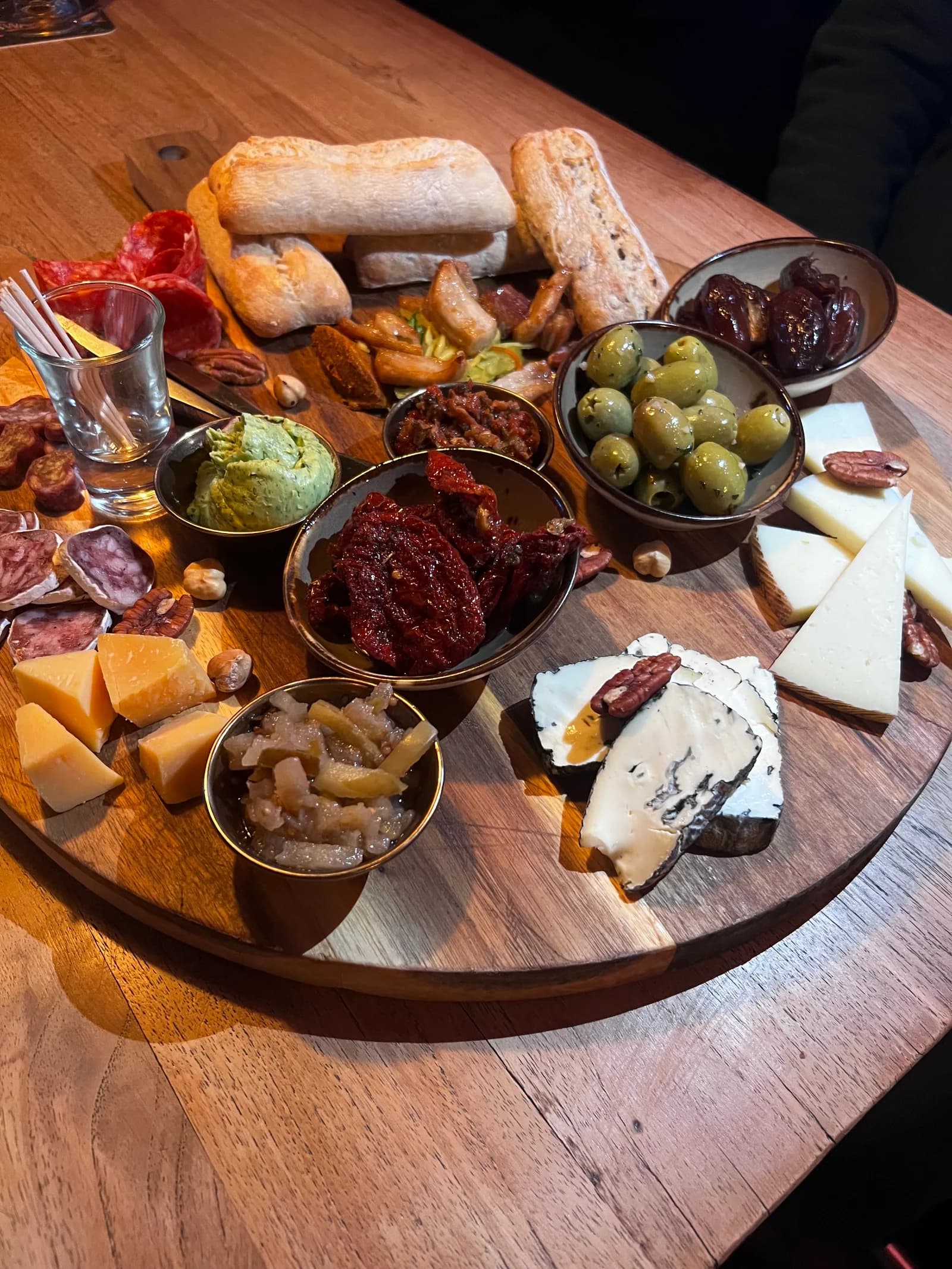 Ronde plank met kazen, charcuterie, olijven, zongedroogde tomaat en stokbrood, met dipschaaltjes ertussen