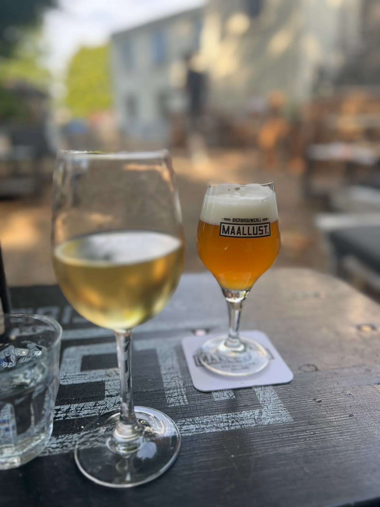 Glas Maallust-bier met logo naast een glas witte wijn op een terrastafel in de zon