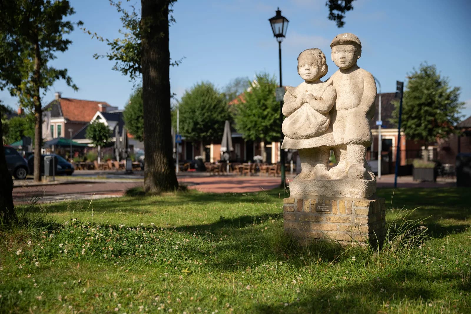 Het stenen beeld van Ot en Sien in het gras op de Brink, met de terrassen van Roden erachter
