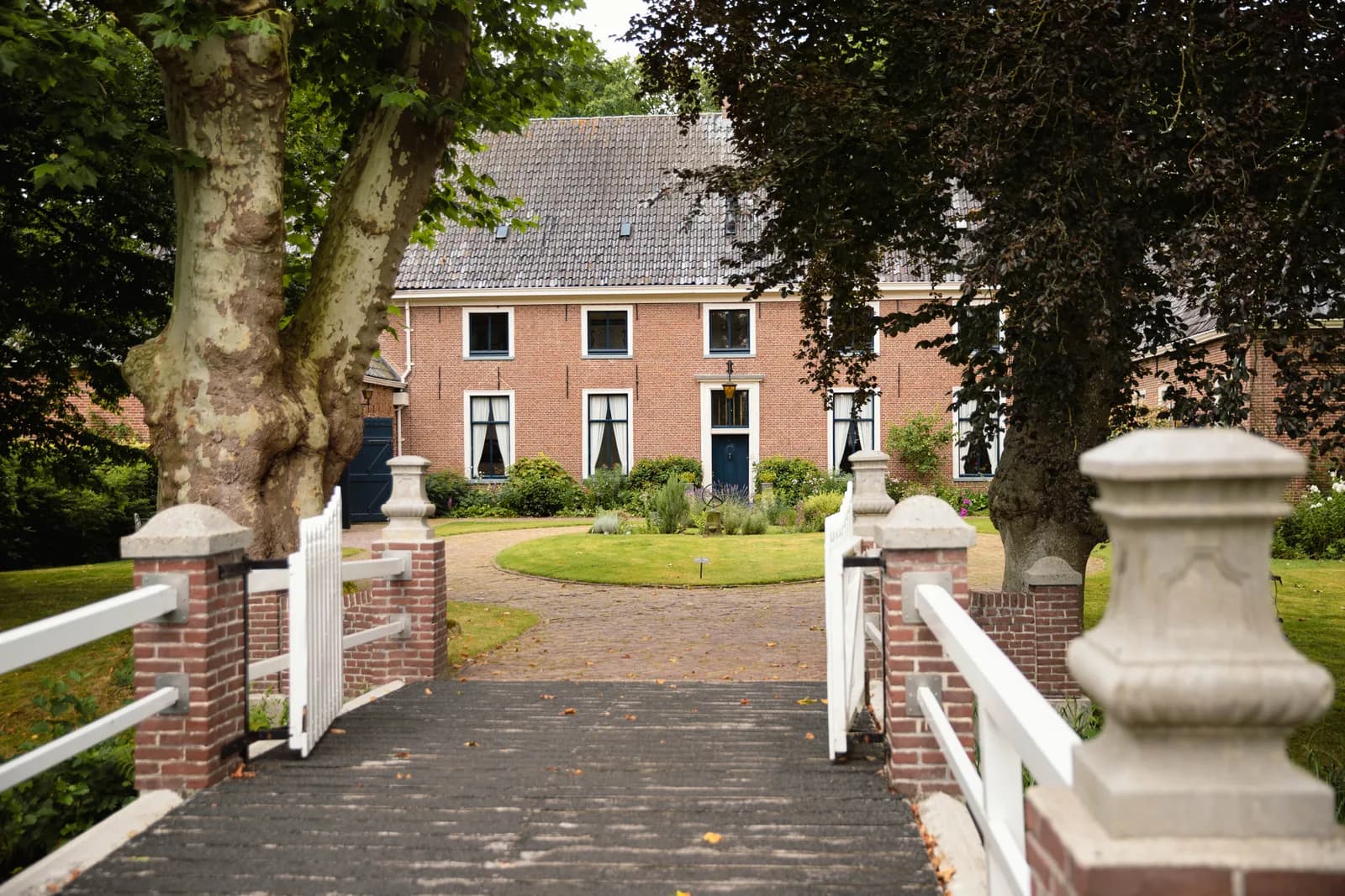Witte brug met bakstenen pilaren over de gracht naar een landhuis met voortuin