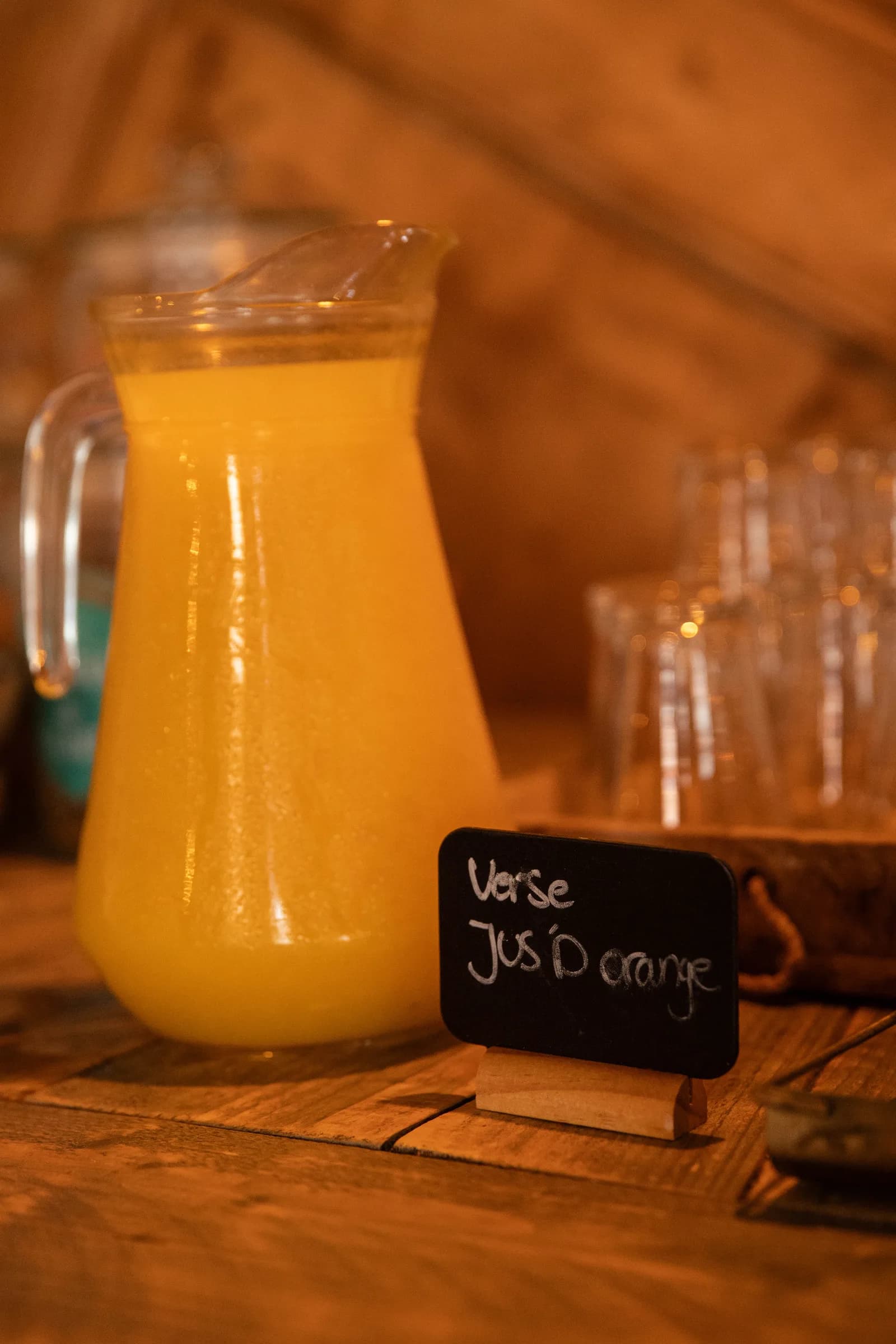Glazen kan verse jus d’orange met een krijtbordje ernaast en glazen op de achtergrond