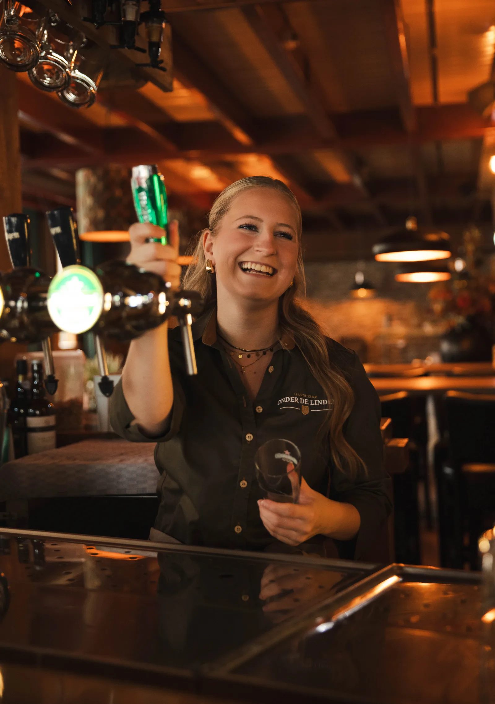 Lachende medewerkster met haar hand aan de biertap en een glas in de andere hand