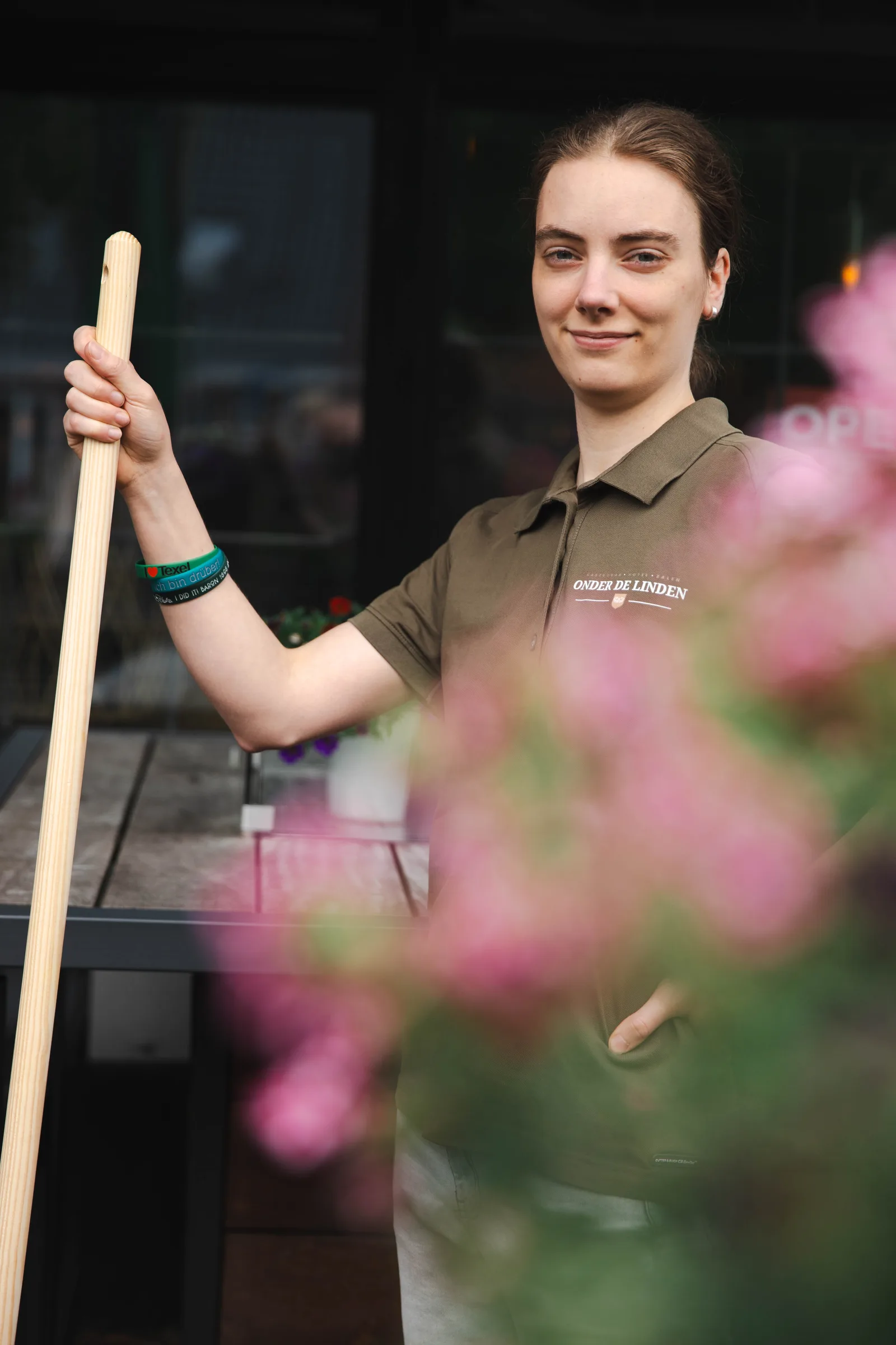 Medewerkster met een bezem in de hand bij de tafels op het terras, met roze bloemen op de voorgrond