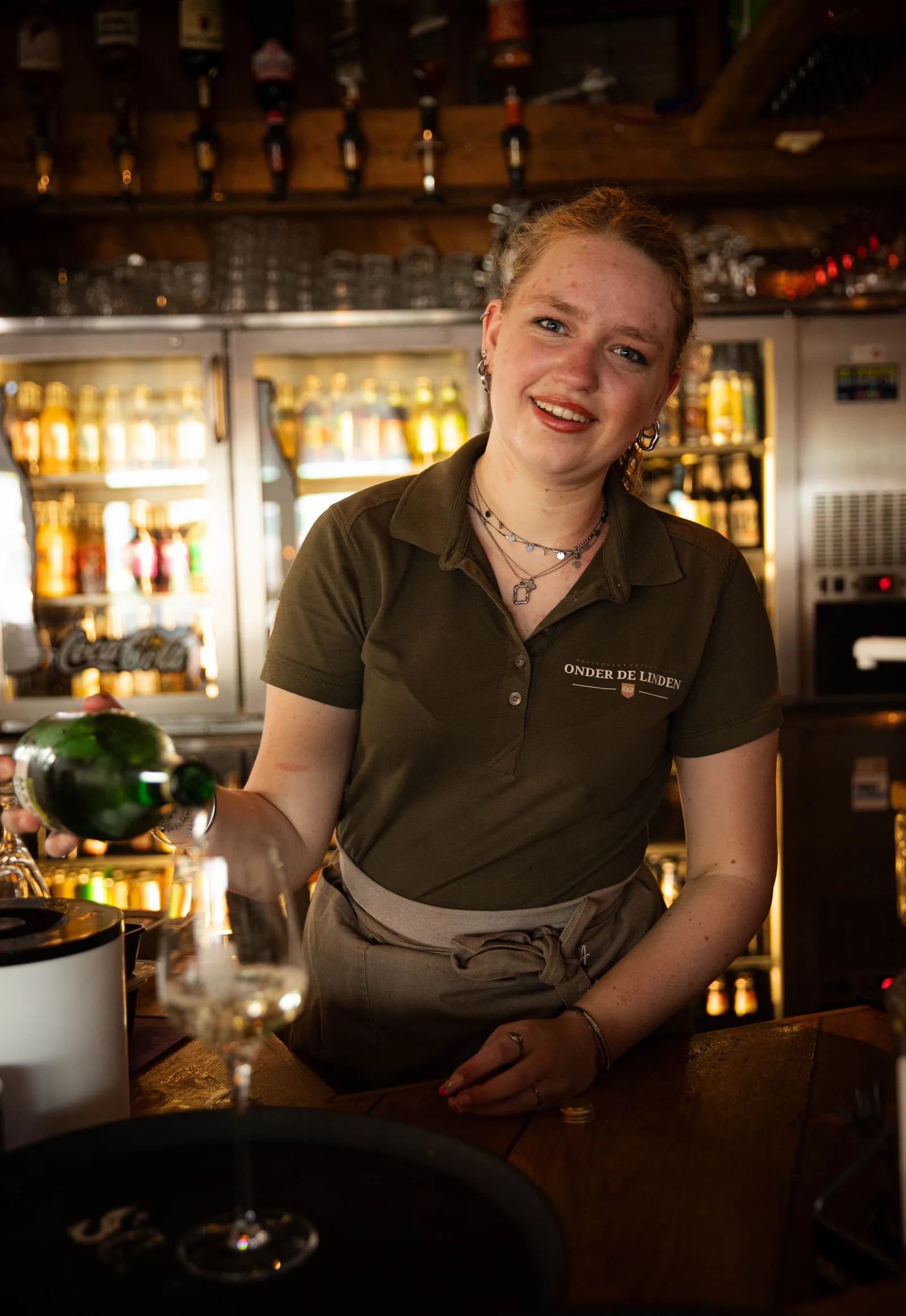 Lachende medewerkster in olijfgroene polo schenkt witte wijn in achter de bar