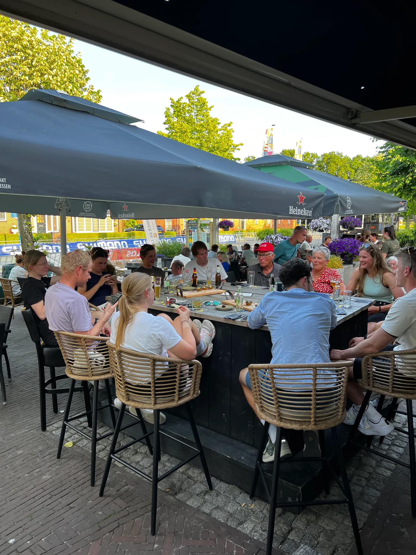 Gasten zitten rondom de buitenbar op het terras onder de parasols, met de Brink erachter