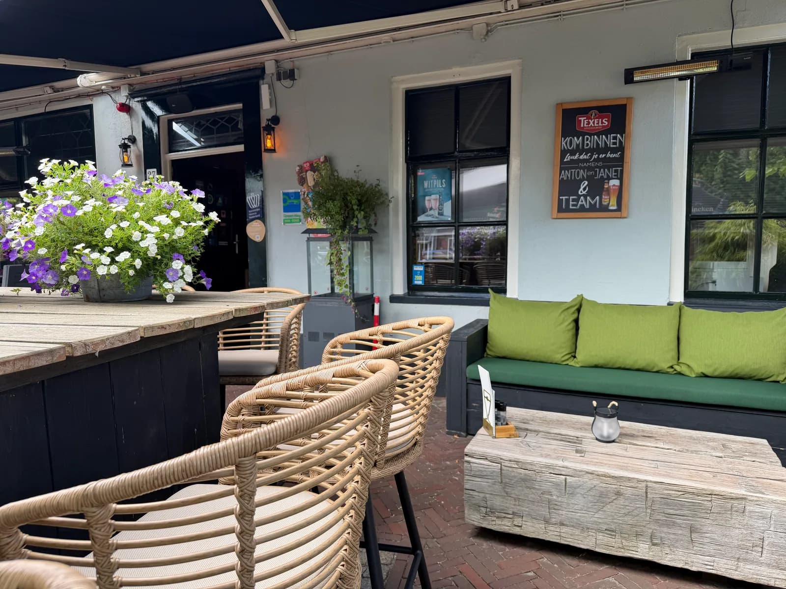 Groene loungebank met kussens onder het krijtbord Kom binnen, met bloembak en rieten stoelen ervoor