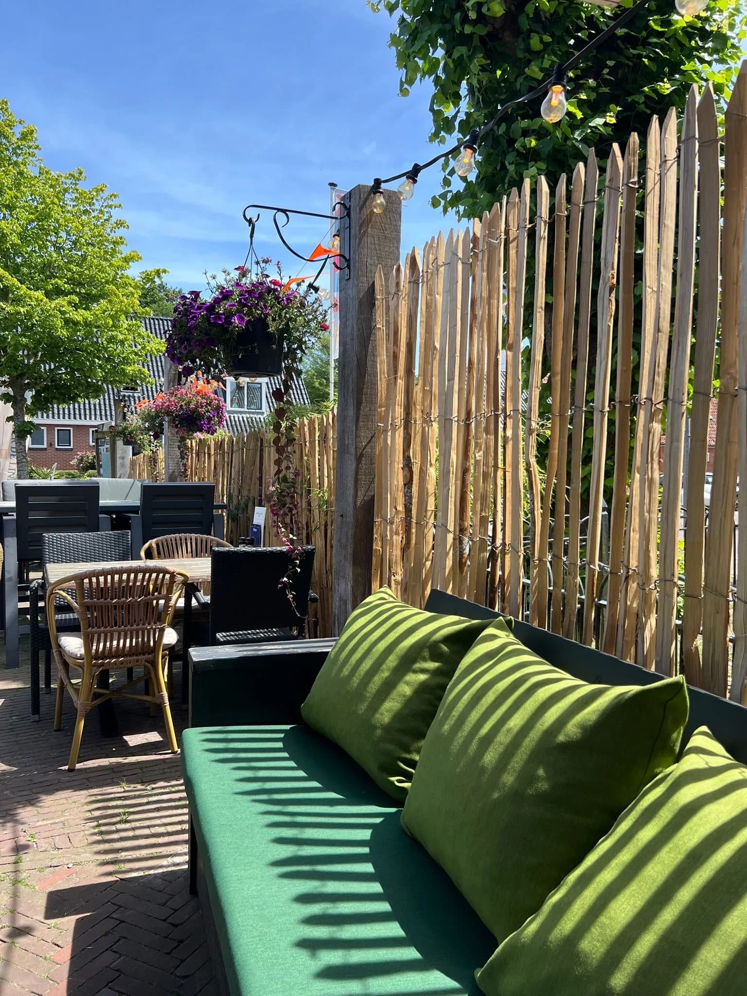 Loungebank met groene kussens langs de houten schutting, met hangbloemen en een lichtsnoer in de zon