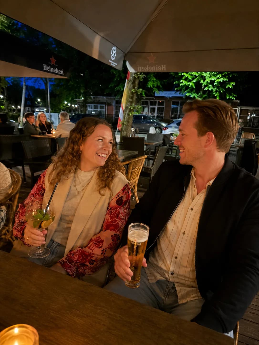 Stel lacht met een bier en een cocktail aan de terrastafel onder de parasol, in de avond
