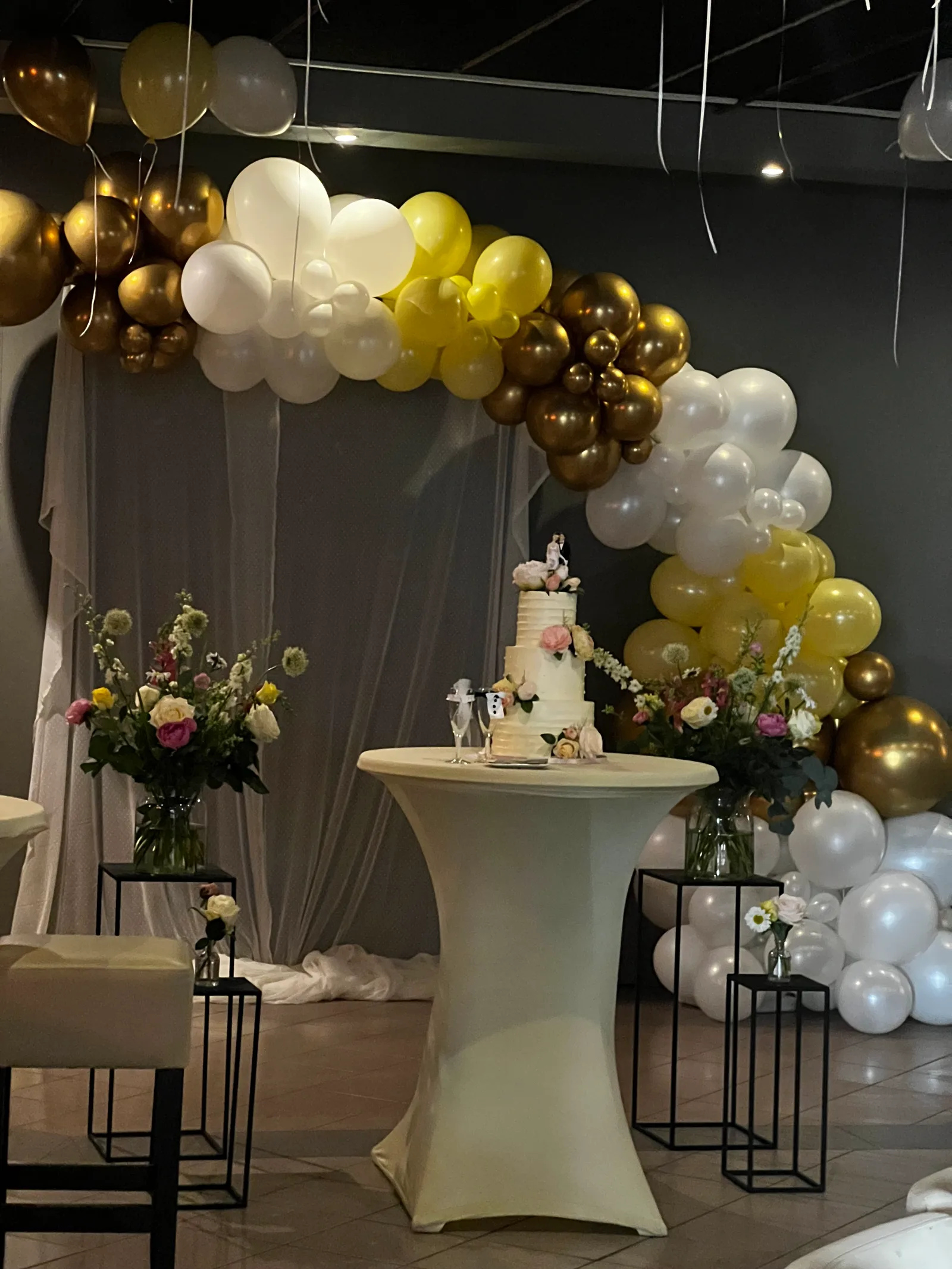 Bruidstaart op een statafel onder een ballonboog in geel, wit en goud, met boeketten ernaast