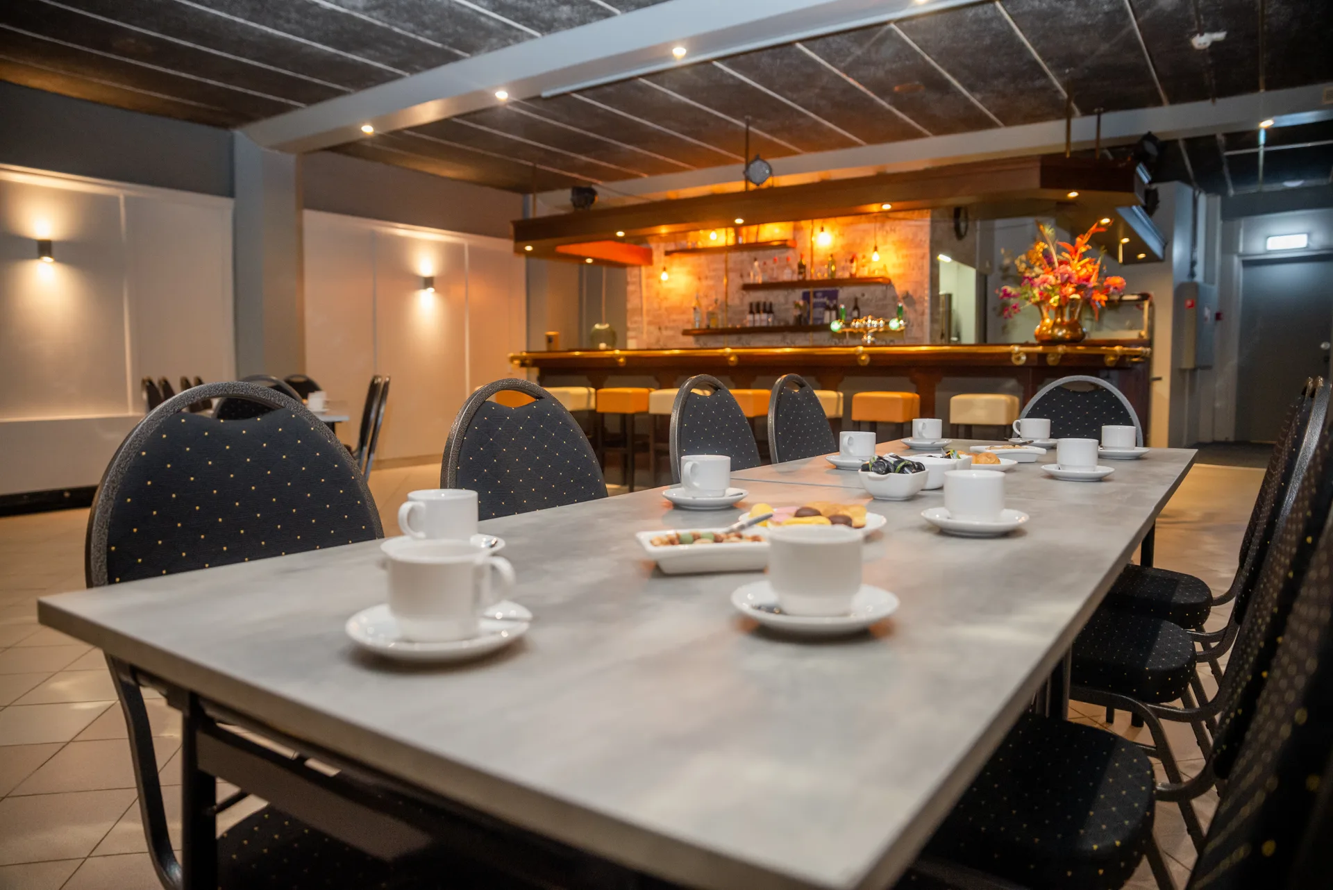Zaal met een lange gedekte tafel, kopjes koffie en de eigen bar op de achtergrond